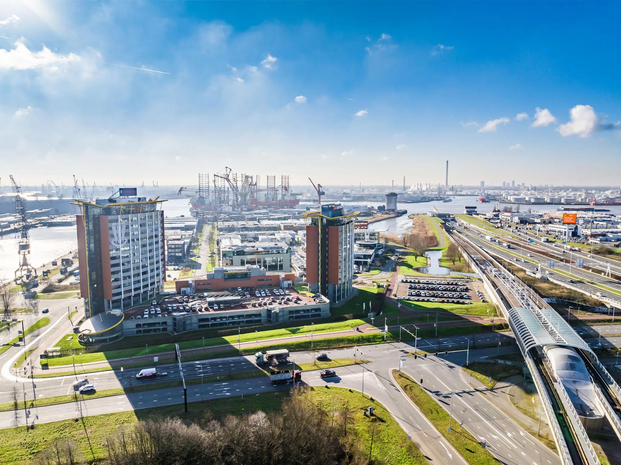 Luchtfoto van de Karel Doormanweg in Rotterdam met kantoorgebouwen, snelweg en havenkranen op de achtergrond.
